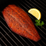 Smoked Pacific rockfish fillet resting on a black grill grate, caramelized with a sweet smoky glaze, garnished with lemon and parsley.